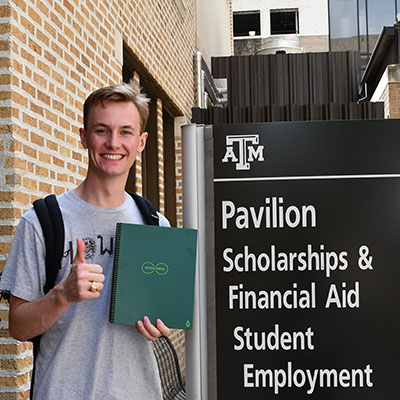 A student (Thomas Jistel) holding a Rocketbook next to the Scholarships and Financial Aid sign.